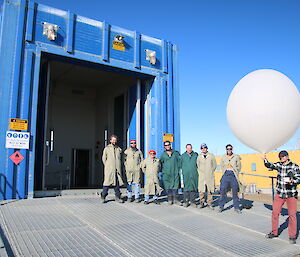 Expeditioners stand outside of a shipping container office, one holds a large balloon