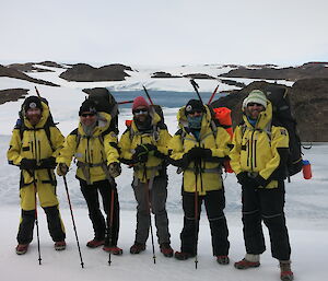 Five expeditioners posing on blue ice with rocky knolls of the Vestfold Hills behind them