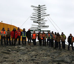 The sixteen current winterers at Davis in front of the signpost outside the Operations building at Davis station