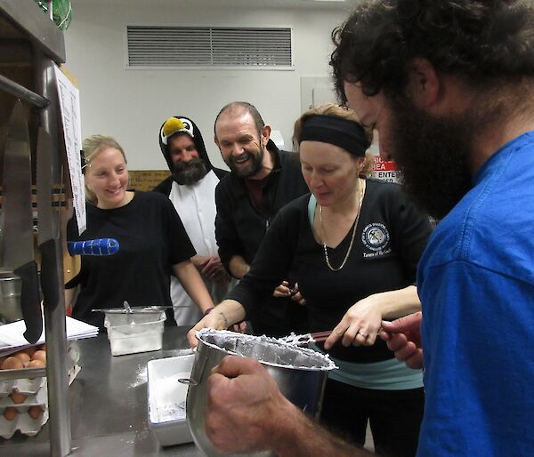 Two female and three male expeditioners making marshmallows in a stainless steel commercial kitchen. One man is dressed as a penguin.