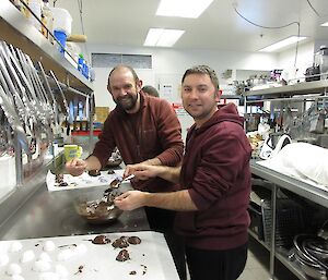 Two male expeditioners in a commercial kitchen coating egg shaped marshmallow in chocolate