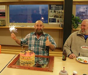 Darren White, who has a long beard and is wearing a flannel shirt, holding sparklers with his pink iced double sponge Birthday Cake
