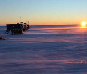 Wind blowing snow low on the ground around the infrastructure at Woop Woop as the sun skims the horizon before setting again