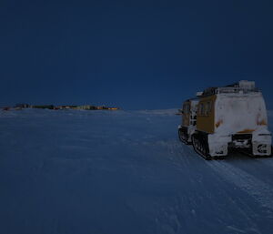 A view of the yellow Hagglund on the sea ice with Davis station in the background, the sunset colour reflecting in the Davis Station windows