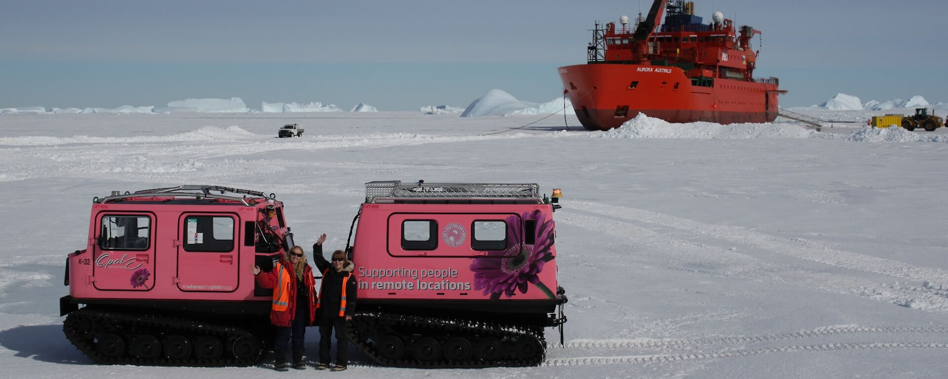Ali and Kirsten wave to us standing in front of the pink Hagg, Opal, which is parked on the sea-ice having been taken off the main sea-ice road and onto the ungroomed sea-ice to see how she handles. In the background is the Aurora Australis parked in the sea-ice surrounded by utes, trucks and other Haggs loading up with supplies being unloaded from the ship during re-supply.