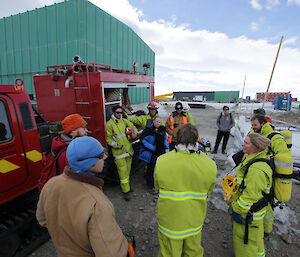 Eleven people are standing in a circle beside the fire truck Hagglund, having a de-brief on how the fire drill went. There are three field training officers, one doctor (Ralph), one Station Leader (Kirsten) and the full fire team, still dressed in their fire fire fighting equipment. Everyone is concentrating on what is being said but looking relaxed as the drill went well..