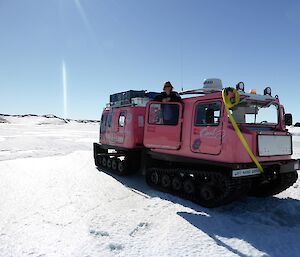 An expeditioner is standing up in the Hägglunds cab. The pink Hägglund is parked on the sea ice. In the distance are islands frozen in by the sea.