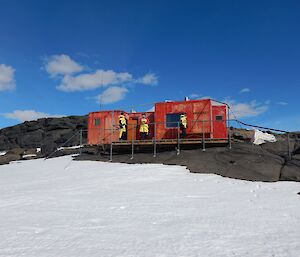 Brookes Hut — a red field hut, nestled in the black rocks of the Vestfold Hills, with a deck out the front, to sit our and enjoy the views. People can be seen on the deck in their bright yellow outdoor gear.