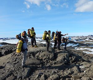 Six people with an FTO, standing on a hill, pointing to a site. There are hills, frozen lakes and Long Fjord in the background. Everyone is carrying full backpacks which include sleeping bags, bivvy bags and ice axes.