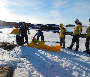 The FTO is unrolling a bivvy bag onto the snow. Four people in their outdoor gear are watching in preparation of having to do this themselves. It is getting later into the night as there are long shadows on the ground.