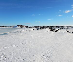 A vista of snow, sea-ice and low lying black rocky hills, with a vast blue sky in sunshine. This is the view from the deck of Brookes Hut, out into Long Fjord.