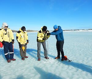 Nick, the field training is showing three expeditioners how to drill a hole in the sea-ice to test the thickness, as a safety measure. They are standing on polished blue sea-ice with the hills of Davis far off in the distance.