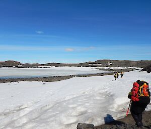 Four people are walking on the snow which runs along the edge of Weddell Lake. They are carrying large backpacks. In the background you can see the frozen lake and low-lying hills.