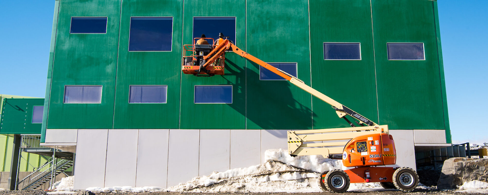 Two tradies are on the elevated work platform, wearing safety gear, cleaning windows on the second storey of the Living Quarters building.