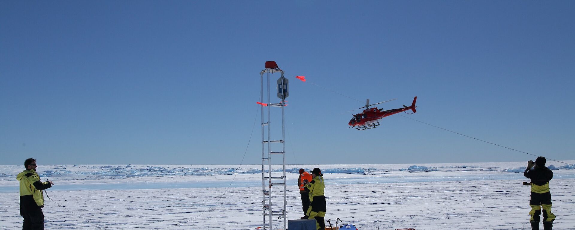 Photo of the tower erected on the glacier, with a helicopter flying overhead, and four people working.