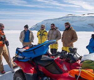 Six people are standing on sea ice, next to a quad bike. The field training officer is holding the drill used to measure sea ice thickness.