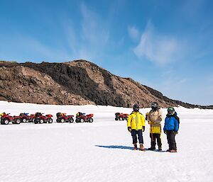 Three people are standing on the sea ice enjoying the view. Behind them is a row of quad bikes and an island.