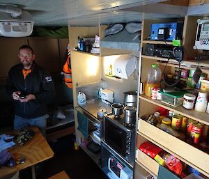 Lance is inside one of the living vans at Woop Woop. There is a tiny kitchen area and a pantry. It looks like the inside of a caravan.