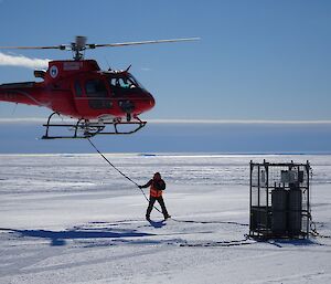 Matt is holding the sling line which is connected to the helicopter hovering above him. He is walking towards a cage which he will connect to the sling line.