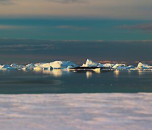 In the foreground is sea-ice, in the middle ground is open water, and in the distance is a line of large, white, icebergs.