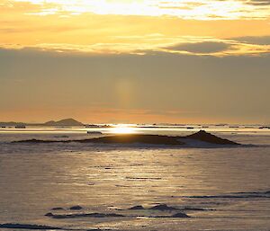 The sun is very low on the horizon, backlighting icebergs on the horizon.