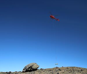 A helicopter is slinging a concrete panel over rocky countryside, carrying it towards the Infrasound worksite.