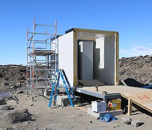 A small hut, still in the process of being built, has a large cabinet sitting in the middle of the floor. The side wall is missing and a ramp is visible. The cabinet looks as tall as the building.