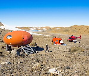 The accommodation at Hop Island: an orange fibre glass hut, known as a smartie as it looks like a smartie on legs. And an apple (a round red fibre glass hut). The helicopter is also seen in the shot.