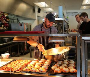 Gideon is putting Hollandaise sauce on egg and bacon muffins in the bay marie. Chefs can be seen in the kitchen behind him.