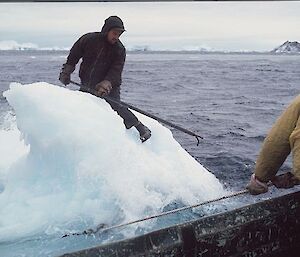 A man is sitting on top of a bergy-bit, he is holding a boating hook. The ice is being put onto a boat to be taken back to station as a source of water.