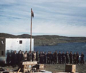 23 men are lined up alongside a building under construction and an Australian flag, for an official opening of station photo.