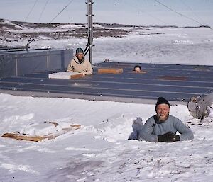 Two men are seen from the waist up. One has popped through the escape hatch on the community hall. The other is half in snow.