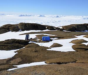 A blue box and some electronic equipment is seen on a rock outcrop, surrounded by plateau ice. It is a seismometer, recording underground vibrations.