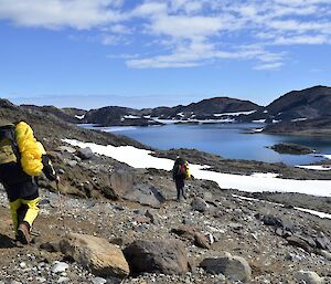 Surveying the geology of the Vestfold Hills.Two people are seen walking down towards a lake.