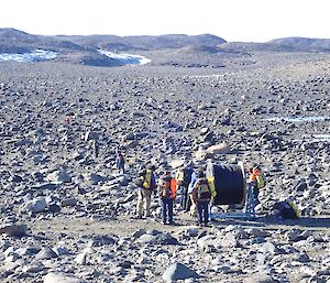An aerial view of ten people in a rocky landscape. Most are standing around a giant spool of cable. A couple are seen “walking” the cable out over the ground.