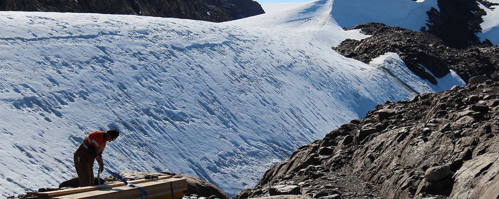 A man is seen unpacking a bundle of long wooden planks. Behind him is the icy Trajer Ridge.