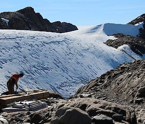 A man is seen unpacking a bundle of long wooden planks. Behind him is the icy Trajer Ridge.