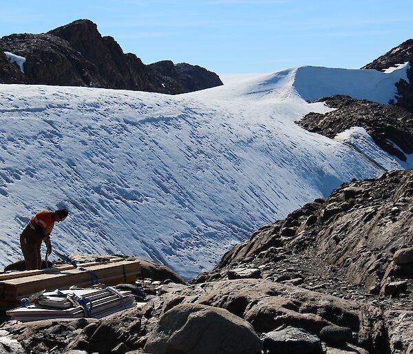 A man is seen unpacking a bundle of long wooden planks. Behind him is the icy Trajer Ridge.