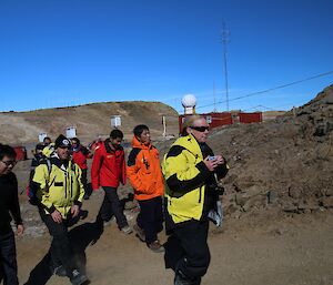 A group of people walk around the station on a tour. Red buildings are seen in the background.