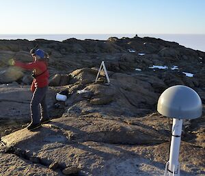 Billy stands on the rock with her arms out, indicating to the helicopter pilot (out of view), where to land. GPS equipment is also seen on site.