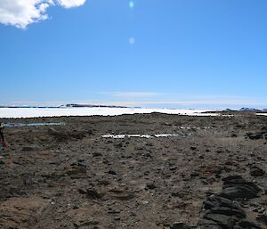 Expeditioner wearing a large backpack and walking along rocky ground, with a frozen fjord in the background.