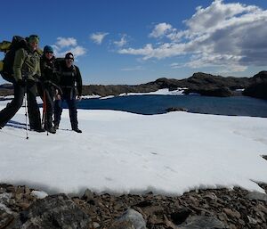 Expeditioners stand on snow in front of a lake, in the rocky Vestfold Hills.