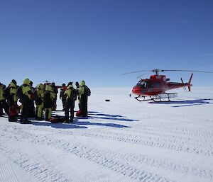 A group of people huddle on the plateau ice, listening to a safety briefing. In the background is the helicopter they are about to use to fly down to Davis Station.