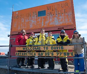 A group of expeditioners arrive at Davis. They are standing behind the sign up by the heli hanger.