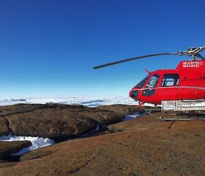 The helicopter is shut down on a rocky area. Behind it the ice of the plateau is visible. The sun is shining and the helicopter is a bright red colour.