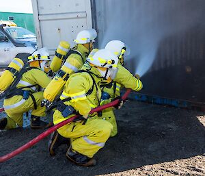 Four people in fire fighting gear and breathing appartus are crouched down in front of an open shipping container. They are putting out a pretend fire in the container.