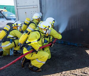 Four people in fire fighting gear and breathing appartus are crouched down in front of an open shipping container. They are putting out a pretend fire in the container.