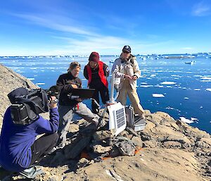 Pete is filming Anna showing Fiona the base station, which is plugged into a computer to download data from cape petrels as to where they have gone to feed. Phoebe looks on. They are on the top of an island with icebergs to sea.