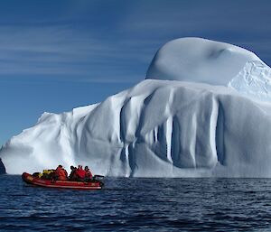 A zodiac travels near an enormous iceberg. The boat looks very small. The iceberg is curvy having rolled over in its history.