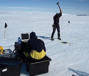 Gideon is seen about to swing a sledge hammer towards a metal plate, sitting on the glacier surface. Tom is sitting on field equipment, collecting data.